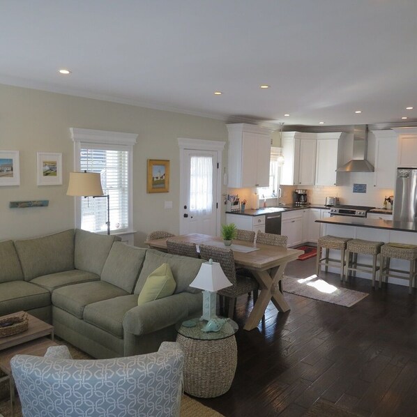 A modern living room with a sectional sofa, a dining table, and a kitchen area with white cabinets and stainless steel appliances.