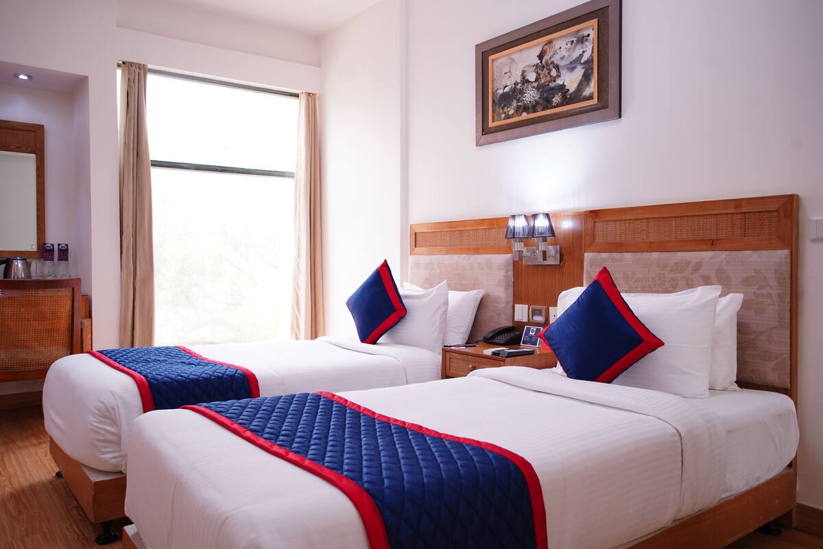 A hotel room with two beds, a wooden headboard, a nightstand, a lamp, and a painting on the wall.