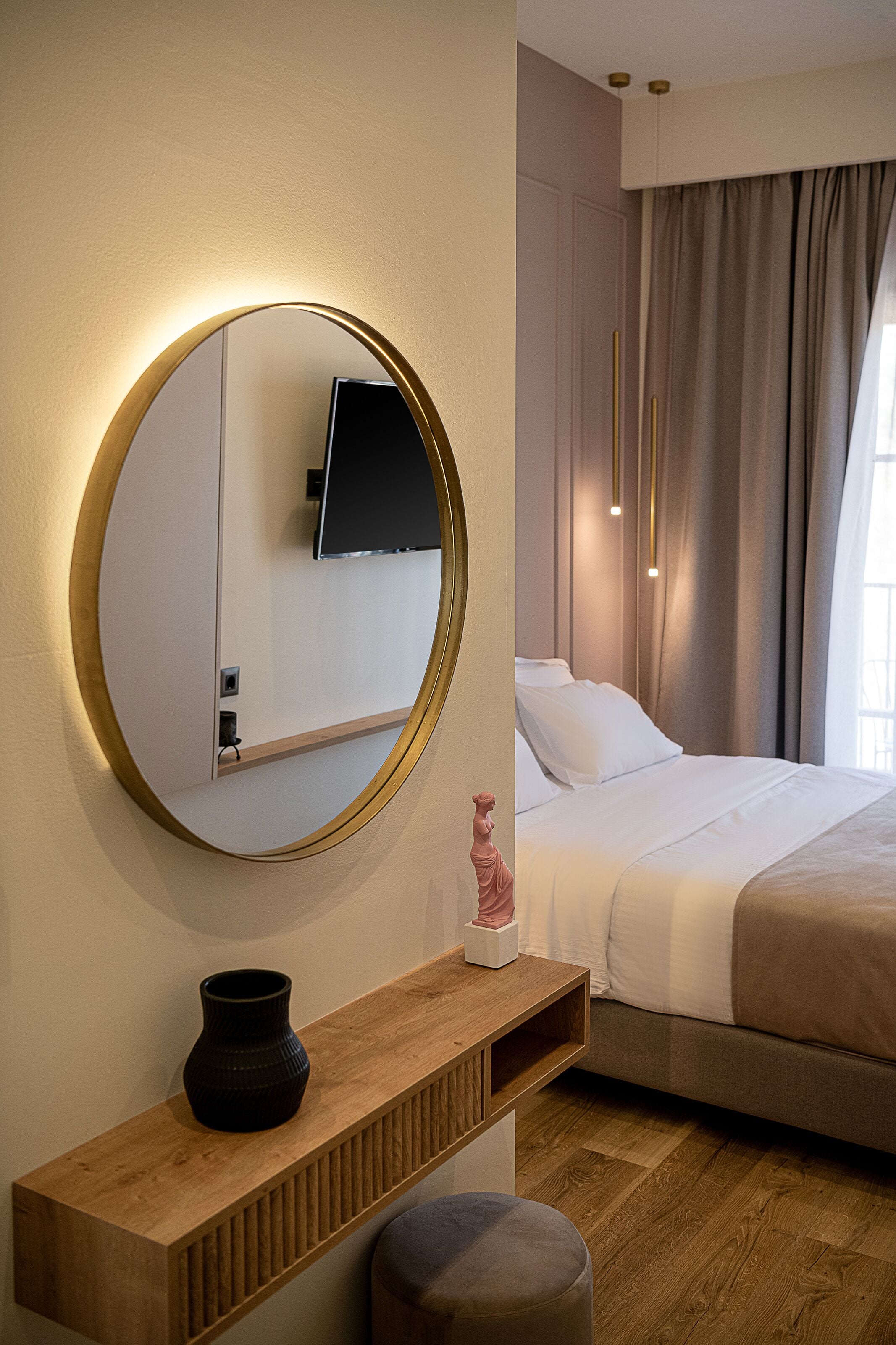 A modern hotel room with a round mirror, a wooden console table, a small statue, and a bed with a beige comforter.