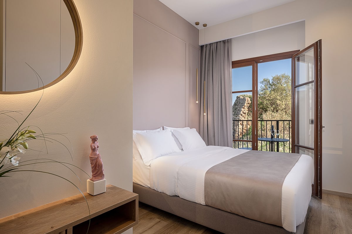 A modern hotel room with a large bed, a bedside table with a vase, a round mirror, and a balcony with a view of trees and rocks.