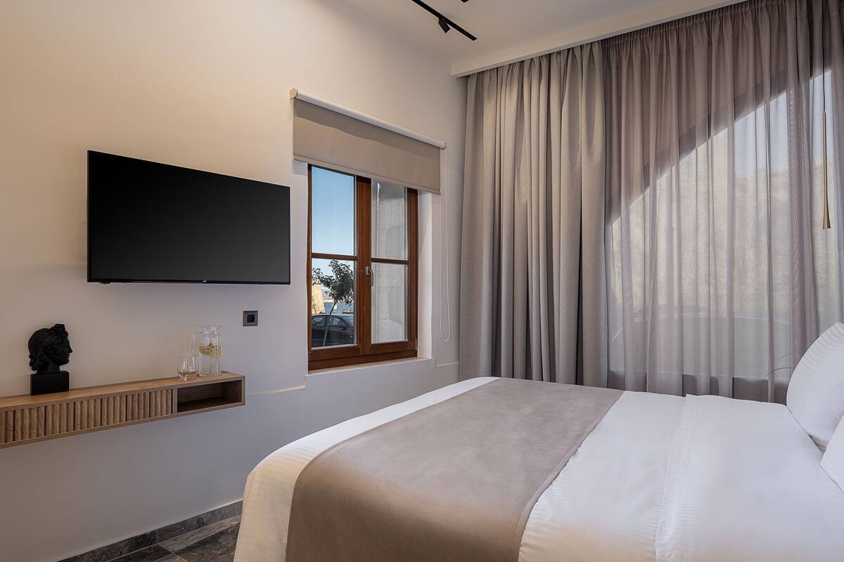 A modern hotel room with a large bed, a flat-screen TV mounted on the wall, a window with a view of the outside, and a built-in shelf with a bust sculpture and a glass.