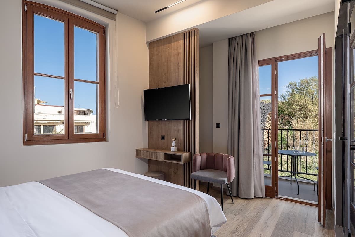 A hotel room with a bed, a TV mounted on the wall, a desk, a chair, and a balcony with a table and chairs.