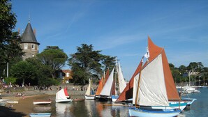 Un port où plusieurs voiliers sont amarrés près du rivage, un bâtiment historique avec une tour en arrière-plan, et des personnes sur la plage.