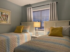 Un dormitorio con dos camas, una mesita de noche con lámpara y una ventana con persianas.