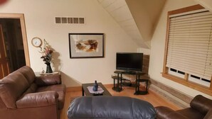 A living room with a brown leather sofa, a black ottoman, a TV on a stand, and a framed picture on the wall. - Tamarack Point Homestead (Arcadia)