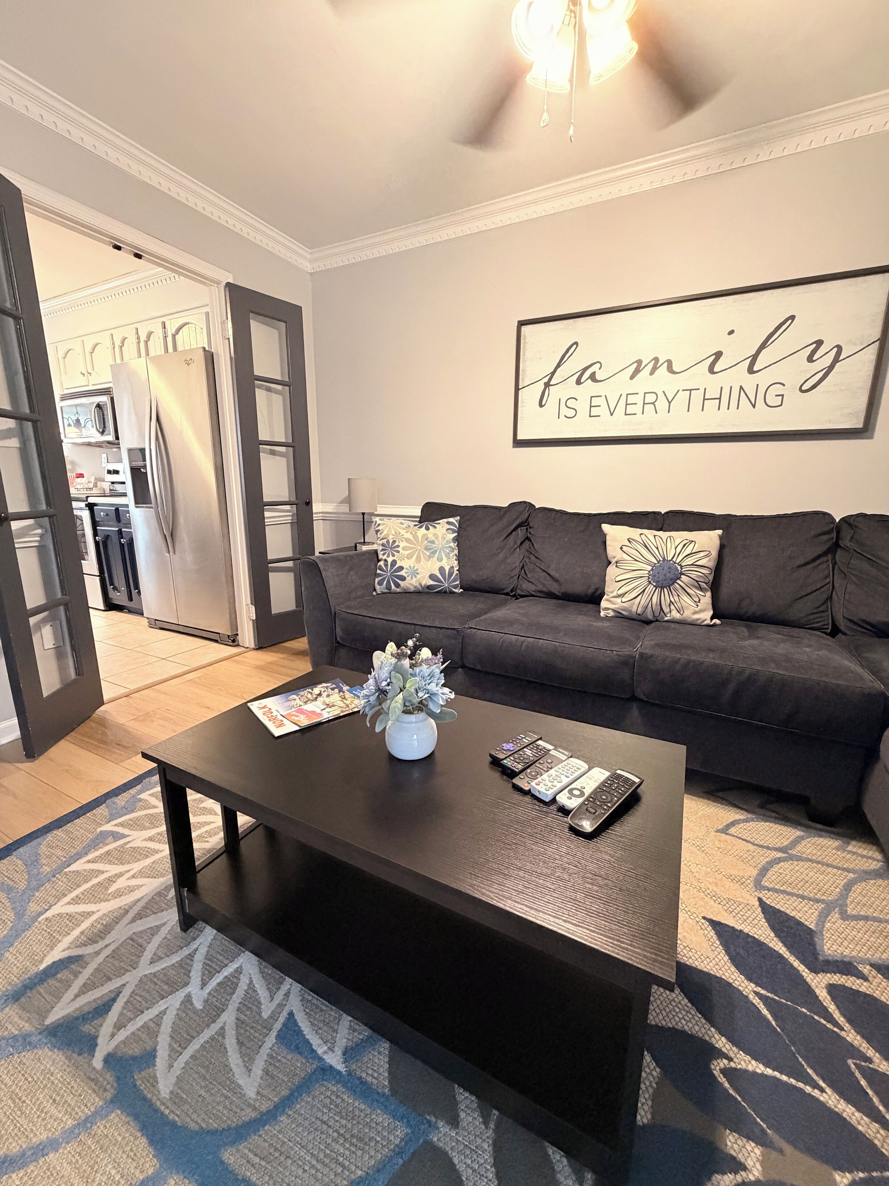 A living room with a dark sofa, a coffee table with a vase and remotes, a kitchen in the background, and a 'family is everything' sign on the wall.