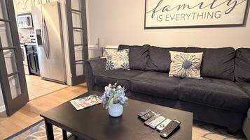 A living room with a dark sofa, a coffee table with a vase and remotes, a kitchen in the background, and a 'family is everything' sign on the wall.