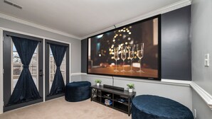 A living room with a large flat-screen TV mounted on the wall, a dark blue sofa, a small side table with a plant, and a sliding glass door with dark curtains.