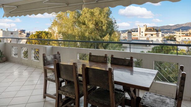 Un balcone con tavolo e sedie, vista su un paesaggio urbano con edifici e alberi.