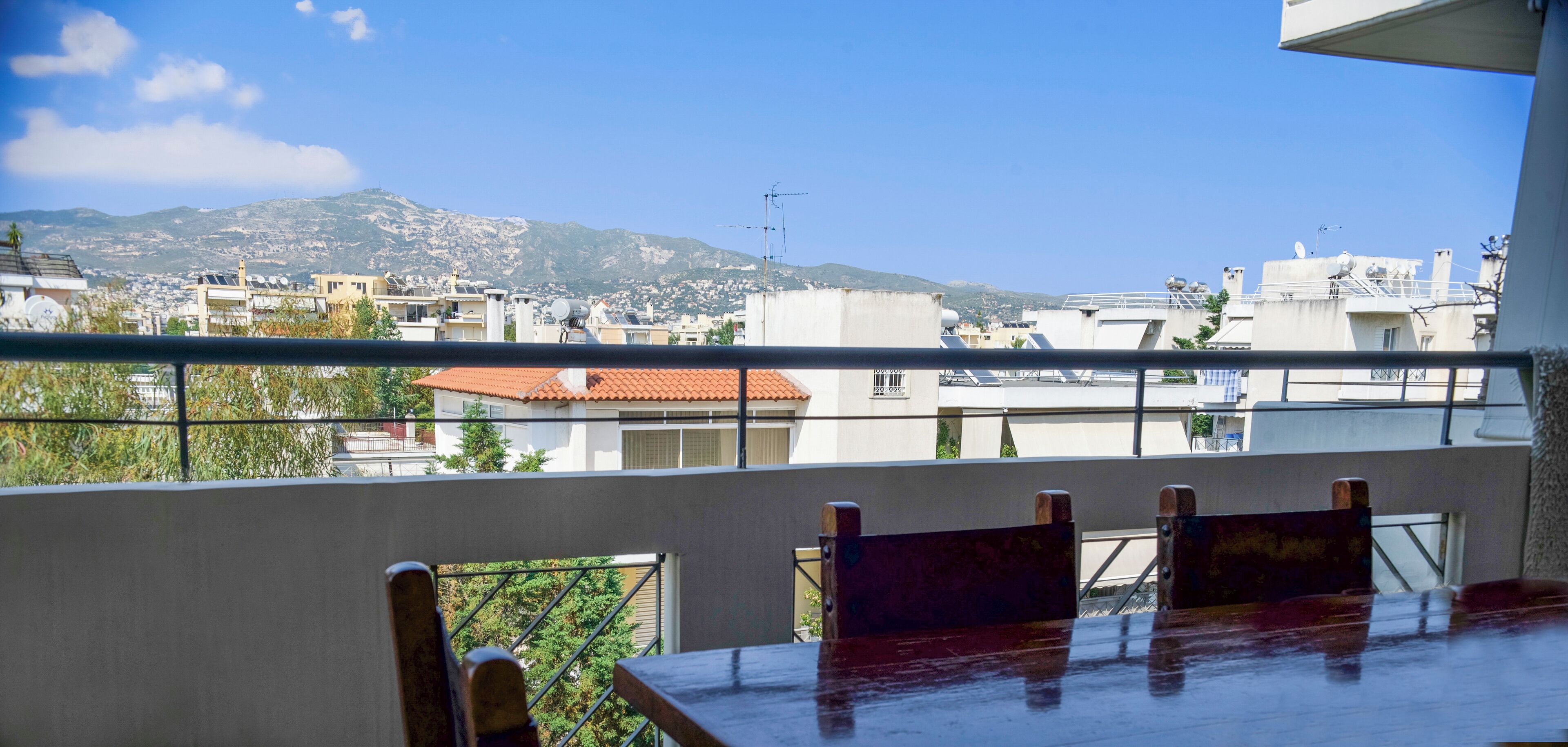A balcony with a table and chairs, overlooking a residential area with buildings and mountains in the background.