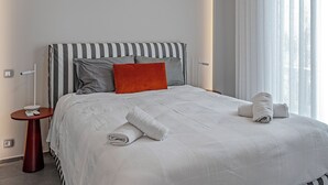 En pænt redt seng med stribet sengetøj, en rød pude og sammenrullede håndklæder i et moderne værelse med et lille rundt bord ved siden af.