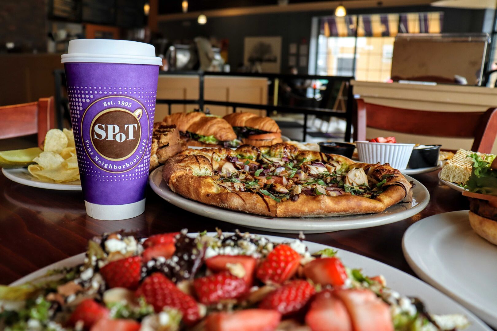 Une table à manger avec une tasse violette portant l’inscription « Café du quartier SPT », une pizza, une assiette de fraises et un sandwich.