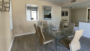A modern dining area with a glass table and metal chairs, a kitchen in the background, and a ceiling light fixture.