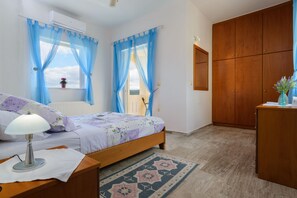 A bedroom with a bed, a nightstand, a desk, a wardrobe, and a window with blue curtains. - Villa Albion (Platanias)