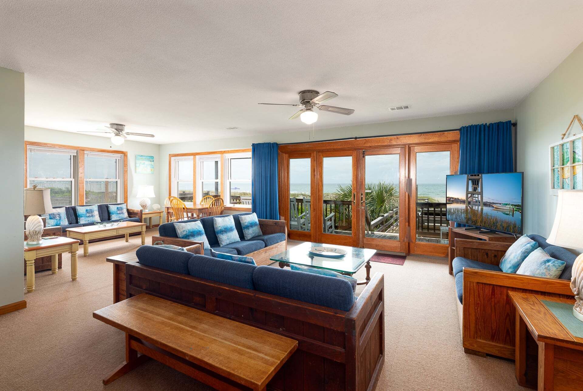 A spacious living area with a large sofa, a coffee table, and a TV. There are multiple windows offering a view of the ocean.
