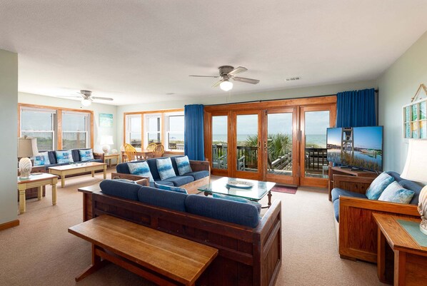 A spacious living area with a large sofa, a coffee table, and a TV. There are multiple windows offering a view of the ocean.