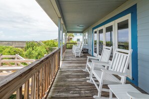 Une terrasse en bois avec des chaises et des tables blanches, donnant sur une plage et l’océan.