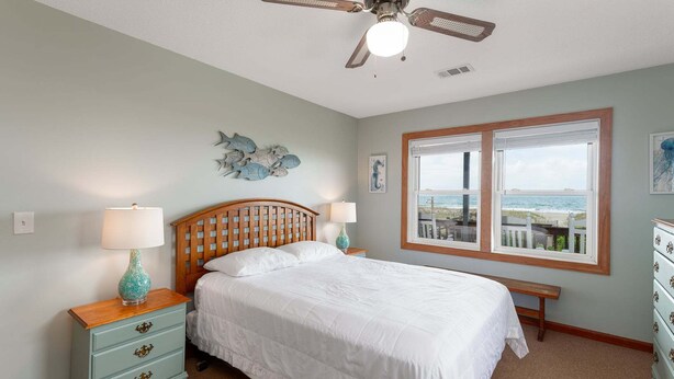 Une chambre à coucher comprenant un lit, une table de chevet, une commode, un ventilateur de plafond et une fenêtre donnant sur la plage.