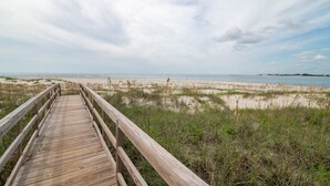 Una pasarela de madera que conduce a una playa con dunas de arena y zonas cubiertas de hierba.