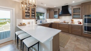 A modern kitchen with a large island, wooden cabinets, stainless steel appliances, and a view of the ocean. - Hobbs Realty - “Shore To Please” | Luxury Oceanfront w/ Saltwater Pool, Hot Tub & Elevator (Holden Beach)
