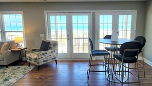 Un salón con sofá, un sillón estampado, una mesa de centro y una mesa de comedor con sillas. Amplias ventanas ofrecen vistas al océano.