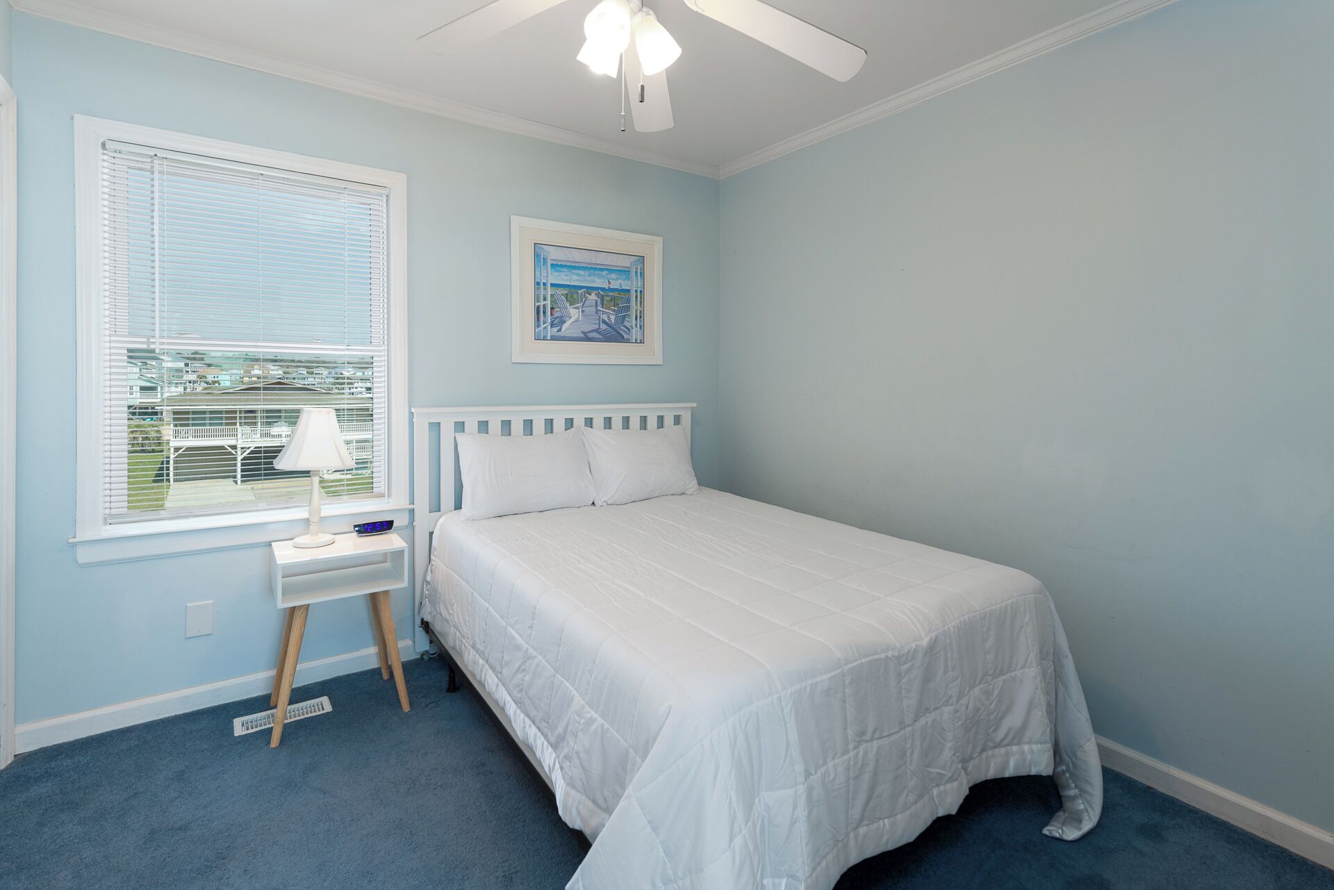 Ein Schlafzimmer mit Bett, Nachttisch, Lampe, Deckenventilator, Fenster mit Jalousien und einem gerahmten Bild an der Wand.