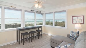 A spacious room with large windows overlooking the beach, a ceiling fan, a bar area with stools, and a cozy seating area with a couch and pillows.