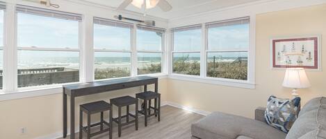 Una habitación amplia con ventanas grandes vistas a la playa, un ventilador de techo, una zona de bar con taburetes y un rincón de descanso con sofá y cojines.