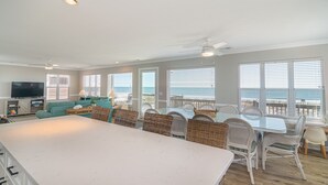 Un vaste espace de vie comprenant une grande cuisine avec un îlot central, une table à manger et des chaises en osier. On y trouve également un téléviseur, un canapé et une vue sur l’océan grâce à de grandes fenêtres.