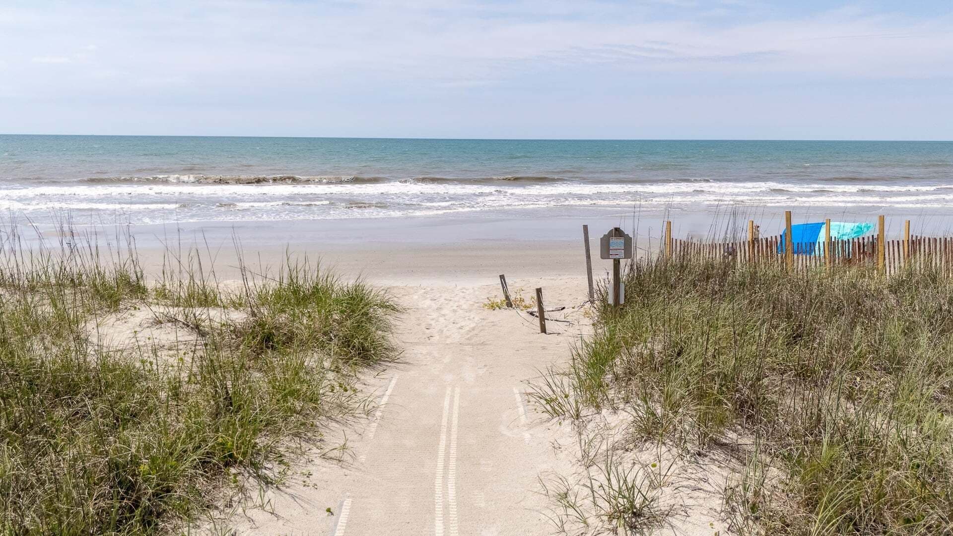 A sandy beach path leading to the ocean, with a wooden fence and a blue structure on the right.