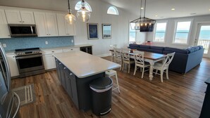 A modern kitchen with white cabinets, a central island, and a dining area with a blue sofa. - Hobbs Realty - Oceanfront home w/Private Pool, Luggage Lift, and Hot Tub (Holden Beach)
