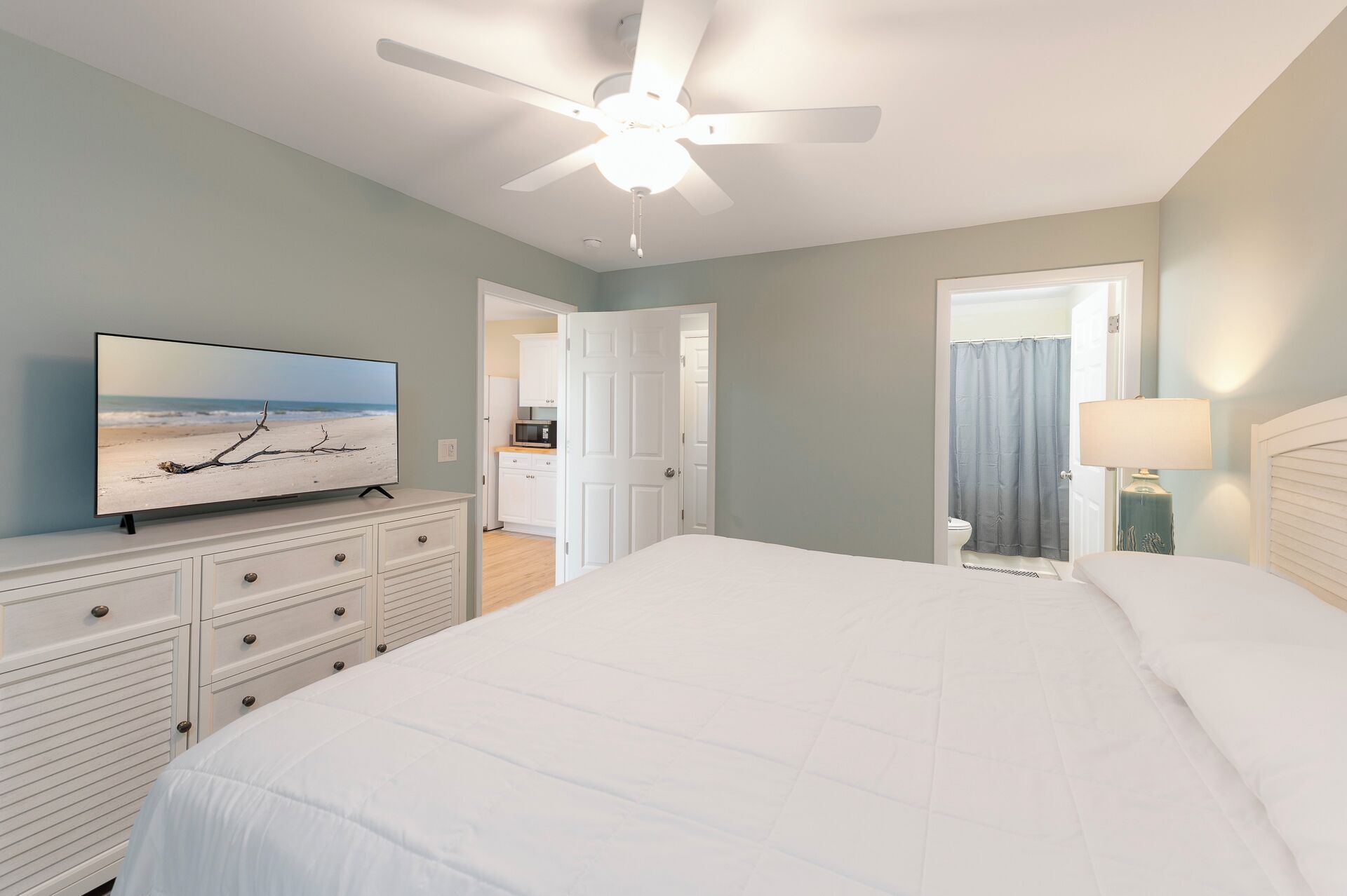 A bedroom with a bed, a TV on a dresser, a ceiling fan, and a bathroom visible through an open door.