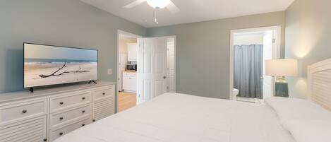 A bedroom with a bed, a TV on a dresser, a ceiling fan, and a bathroom visible through an open door.