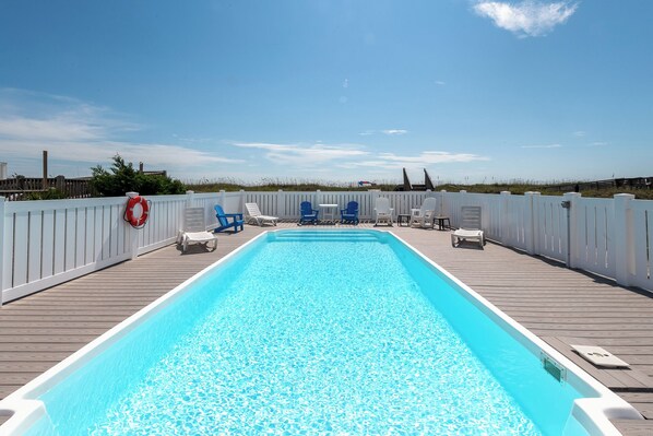 Una piscina rectangular con cubierta de madera, rodeada de vallas blancas y sillones de descanso azules y blancos.