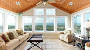 A spacious room with a high ceiling, large windows offering ocean views, a ceiling fan, and a modern coffee table.