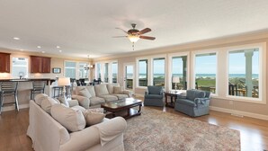 A spacious living room with a large sofa, a coffee table, and a view of the ocean through large windows. - Hobbs Realty - "That's It II" an amazing oceanfront home with lovely views, pool, and an elevator. (Holden Beach)