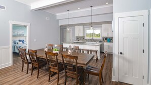 Un comedor amplio con una gran mesa de madera y sillas, una cocina con armarios blancos y un ventanal, y una puerta que da a otra habitación.