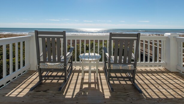A wooden deck with two rocking chairs and a small table, overlooking a beach with a clear view of the ocean. - Hobbs Realty - Oceanfront Dog Friendly w/Private Pool & EV Charger (Holden Beach)
