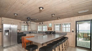 Cucina con mobili in legno, frigorifero in acciaio inossidabile, una grande isola con sgabelli da bar e vista su una terrazza esterna attraverso una porta in vetro.