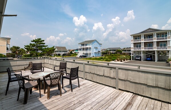 Una terraza de madera con mobiliario exterior, vista a casas frente al mar y cielo despejado.
