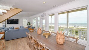 A spacious living area with a large wooden dining table, wicker chairs, a grey sofa, and a view of the beach through large windows.