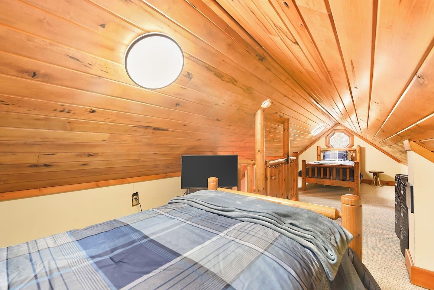Un dormitorio acogedor en el ático con techo de madera, un tragaluz redondo, una cama con colcha de cuadros, un televisor y una mesita pequeña.