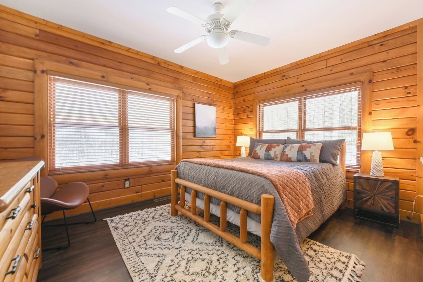 Un dormitorio de cabaña de madera con una cama, dos mesitas de noche, una silla y un ventilador de techo.