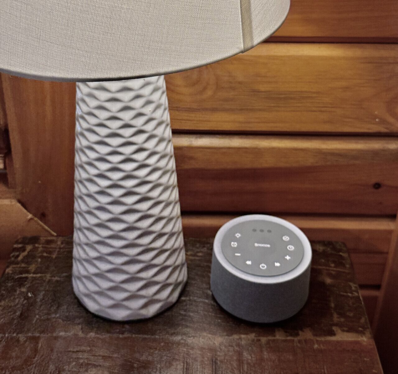 Una lámpara con textura, pantalla circular y un pequeño dispositivo redondo gris con botones sobre una superficie de madera.
