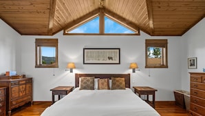Une chambre avec un plafond en bois, un grand lit, deux tables de chevet avec des lampes, une commode et un tableau encadré au mur.