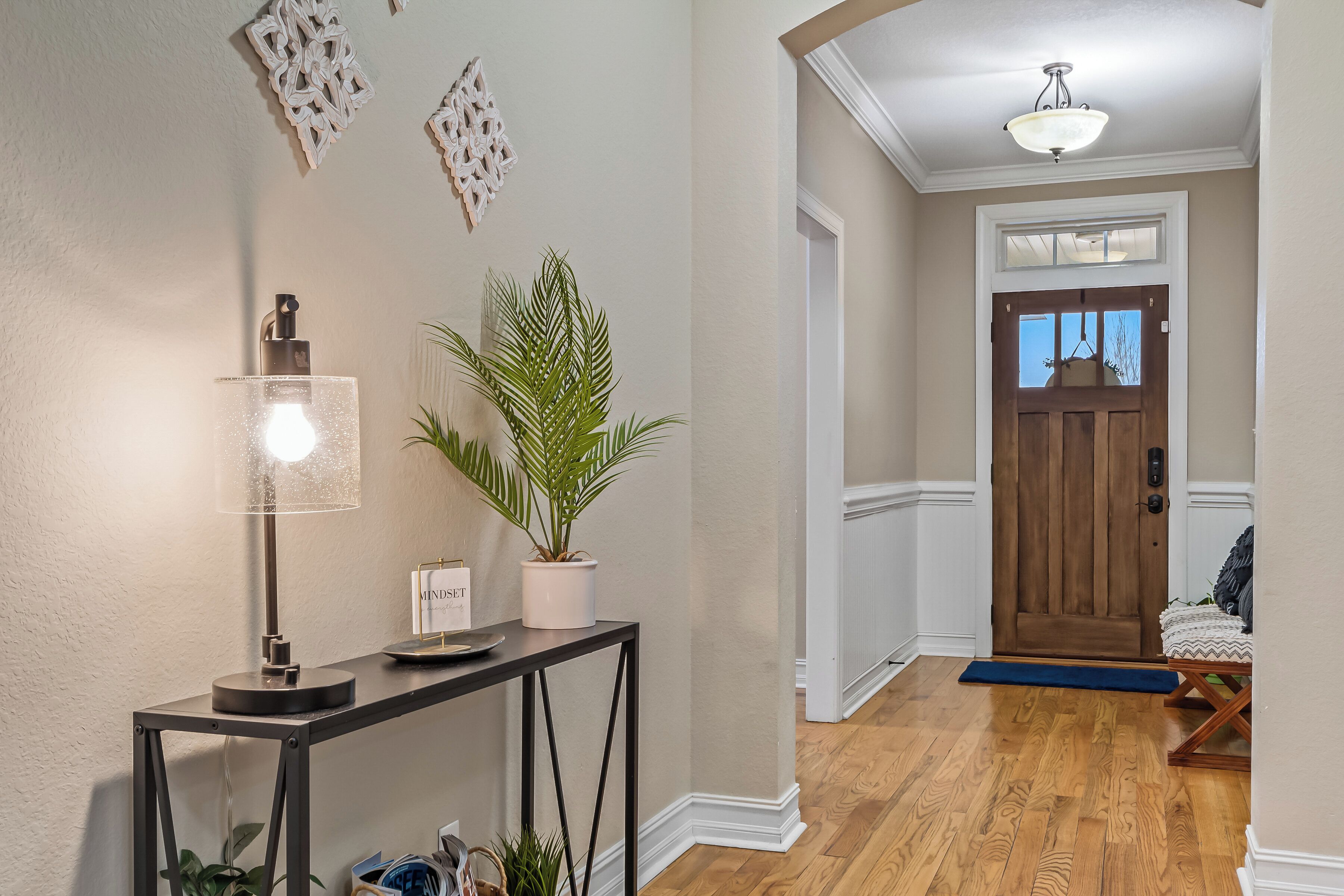 A hallway with a wooden floor, a black console table with a lamp, a potted plant, and decorative wall art.