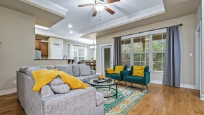 A living room with a gray sofa, a green armchair, and a coffee table. There is a ceiling fan and a window with curtains. - Manassas House 4 BDR  Patio Firepit (Tallahassee)