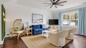 Un salon moderne avec un canapé bleu, une table basse blanche et des fauteuils à motifs. Un ventilateur de plafond et un téléviseur sont fixés au mur.