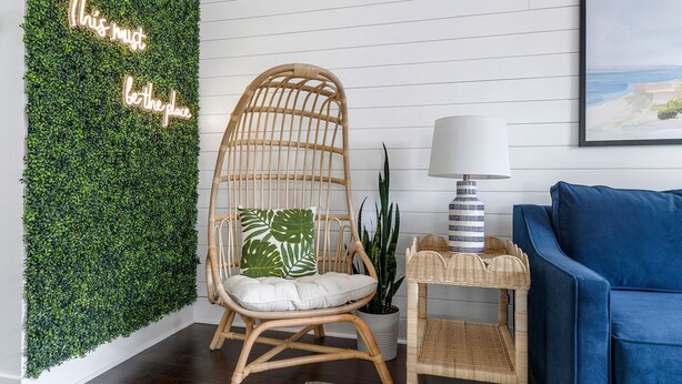 Une pièce avec un mur vert, une chaise en osier, une petite table d’appoint, un canapé bleu et un tableau encadré.