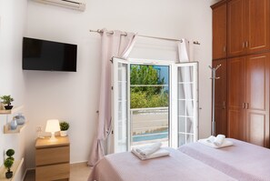 Une chambre avec une télévision, une lampe de chevet, une fenêtre avec des rideaux, un lit avec des serviettes, une armoire en bois et une petite plante sur une table de chevet.